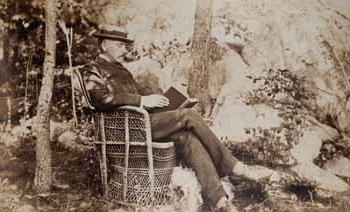 James Lowndes at Beverly Farms, seated outdoors in wicker chair, reading book, with dog at feet Photograph